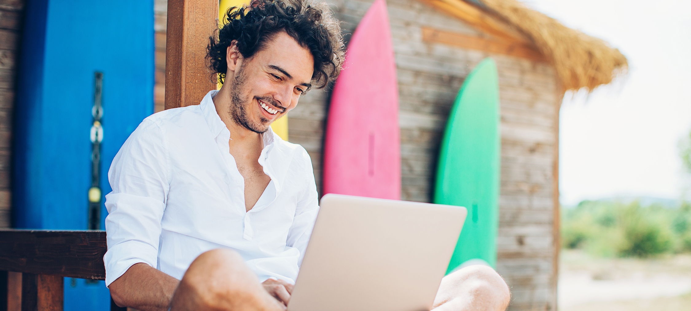 Cheerful young man using laptop with surfing boards at the background.