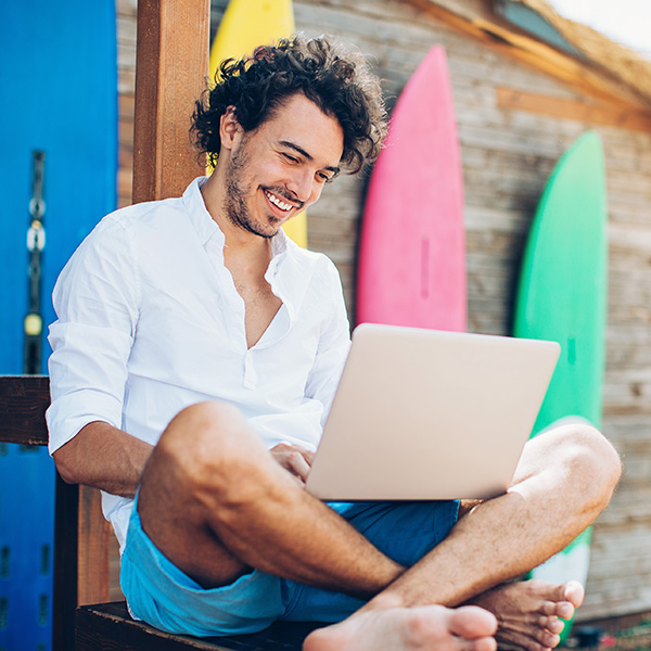 Cheerful young man using laptop with surfing boards at the background.